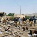 A wide-angle photograph capturing the aftermath of a suicide attack on a security checkpoint in Dera Ismail Khan, Pakistan. Uniformed police officers and civilians walk amidst widespread destruction, with rubble, scattered documents, and broken furniture covering the ground. A damaged tanker and power pylon stand in the background, and a Urdu text banner at the bottom reports multiple officials martyred.