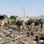 A wide-angle photograph capturing the aftermath of a suicide attack on a security checkpoint in Dera Ismail Khan, Pakistan. Uniformed police officers and civilians walk amidst widespread destruction, with rubble, scattered documents, and broken furniture covering the ground. A damaged tanker and power pylon stand in the background, and a Urdu text banner at the bottom reports multiple officials martyred.