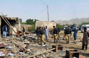 A wide-angle photograph capturing the aftermath of a suicide attack on a security checkpoint in Dera Ismail Khan, Pakistan. Uniformed police officers and civilians walk amidst widespread destruction, with rubble, scattered documents, and broken furniture covering the ground. A damaged tanker and power pylon stand in the background, and a Urdu text banner at the bottom reports multiple officials martyred.