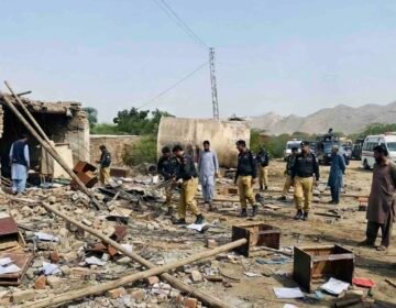 A wide-angle photograph capturing the aftermath of a suicide attack on a security checkpoint in Dera Ismail Khan, Pakistan. Uniformed police officers and civilians walk amidst widespread destruction, with rubble, scattered documents, and broken furniture covering the ground. A damaged tanker and power pylon stand in the background, and a Urdu text banner at the bottom reports multiple officials martyred.