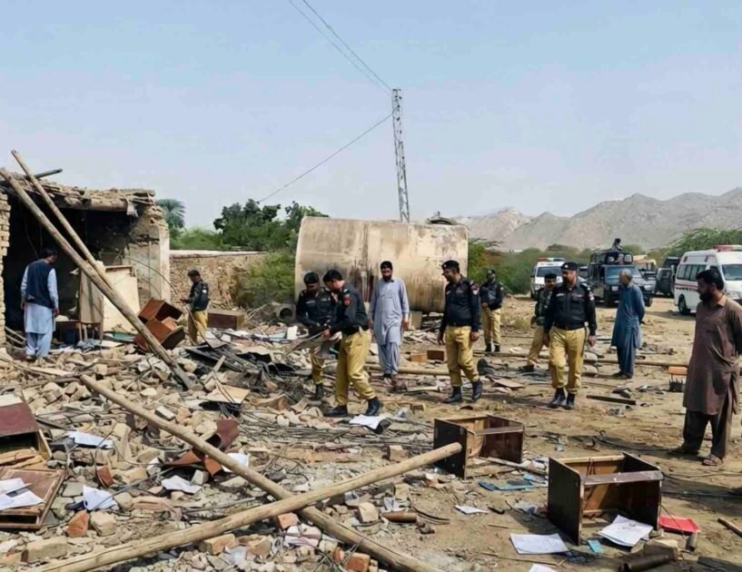 A wide-angle photograph capturing the aftermath of a suicide attack on a security checkpoint in Dera Ismail Khan, Pakistan. Uniformed police officers and civilians walk amidst widespread destruction, with rubble, scattered documents, and broken furniture covering the ground. A damaged tanker and power pylon stand in the background, and a Urdu text banner at the bottom reports multiple officials martyred.