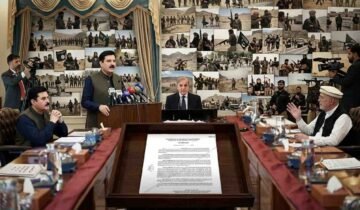 A wide-angle, hyper-detailed photograph capturing an official cabinet meeting in a grand, somber hall in Peshawar. Pakistani Prime Minister Mian Shehbaz Sharif and other key figures, including the specific men from input photos (one in a cap, one in a green vest, one speaking), are seated around a long table. An official notification from the KP Home and Tribal Affairs Department is legible on the table, detailing Governor's Rule and MPO powers under Articles 232 and 234, dated '21-10-2026'. The central wall background, replacing a portrait, features a massive, complex, and somber photo collage depicting various armed and active jihadist groups and militant activities in training and combat across desert landscapes. Camera crews record. The atmosphere is serious and official.