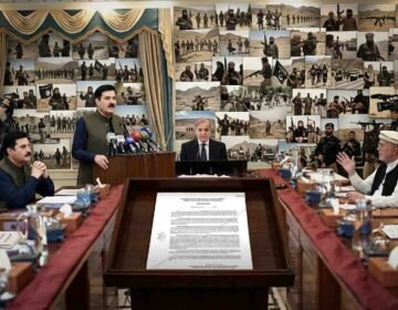 A wide-angle, hyper-detailed photograph capturing an official cabinet meeting in a grand, somber hall in Peshawar. Pakistani Prime Minister Mian Shehbaz Sharif and other key figures, including the specific men from input photos (one in a cap, one in a green vest, one speaking), are seated around a long table. An official notification from the KP Home and Tribal Affairs Department is legible on the table, detailing Governor's Rule and MPO powers under Articles 232 and 234, dated '21-10-2026'. The central wall background, replacing a portrait, features a massive, complex, and somber photo collage depicting various armed and active jihadist groups and militant activities in training and combat across desert landscapes. Camera crews record. The atmosphere is serious and official.