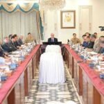 An ultra-high-resolution (8K), wide-angle, professional news photograph depicting a large cabinet meeting in the Prime Minister's House, Islamabad. Prime Minister Mian Shehbaz Sharif is seated at the central head of a long, polished wooden conference table, chairing the meeting. He is in a dark suit, and suited officials are seated on both sides of the table. A prominent military officer is seated near a large framed portrait of Muhammad Ali Jinnah (the Quaid-e-Azam) on the far wall. An elaborate crystal chandelier hangs above the portrait. The unusual cylindrical object covered in textured white velvet textile, centered on the table, is sharply detailed. Two cameramen with professional broadcast cameras are clearly visible, one on the far left and one on the far right. The faces of all dozens of attendees, including a woman in a pink shirt, are crystal clear and distinct. The scene is well-lit and perfectly composed.