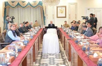 An ultra-high-resolution (8K), wide-angle, professional news photograph depicting a large cabinet meeting in the Prime Minister's House, Islamabad. Prime Minister Mian Shehbaz Sharif is seated at the central head of a long, polished wooden conference table, chairing the meeting. He is in a dark suit, and suited officials are seated on both sides of the table. A prominent military officer is seated near a large framed portrait of Muhammad Ali Jinnah (the Quaid-e-Azam) on the far wall. An elaborate crystal chandelier hangs above the portrait. The unusual cylindrical object covered in textured white velvet textile, centered on the table, is sharply detailed. Two cameramen with professional broadcast cameras are clearly visible, one on the far left and one on the far right. The faces of all dozens of attendees, including a woman in a pink shirt, are crystal clear and distinct. The scene is well-lit and perfectly composed.