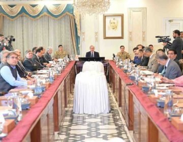 An ultra-high-resolution (8K), wide-angle, professional news photograph depicting a large cabinet meeting in the Prime Minister's House, Islamabad. Prime Minister Mian Shehbaz Sharif is seated at the central head of a long, polished wooden conference table, chairing the meeting. He is in a dark suit, and suited officials are seated on both sides of the table. A prominent military officer is seated near a large framed portrait of Muhammad Ali Jinnah (the Quaid-e-Azam) on the far wall. An elaborate crystal chandelier hangs above the portrait. The unusual cylindrical object covered in textured white velvet textile, centered on the table, is sharply detailed. Two cameramen with professional broadcast cameras are clearly visible, one on the far left and one on the far right. The faces of all dozens of attendees, including a woman in a pink shirt, are crystal clear and distinct. The scene is well-lit and perfectly composed.