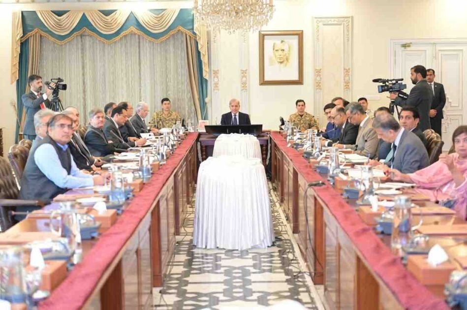 An ultra-high-resolution (8K), wide-angle, professional news photograph depicting a large cabinet meeting in the Prime Minister's House, Islamabad. Prime Minister Mian Shehbaz Sharif is seated at the central head of a long, polished wooden conference table, chairing the meeting. He is in a dark suit, and suited officials are seated on both sides of the table. A prominent military officer is seated near a large framed portrait of Muhammad Ali Jinnah (the Quaid-e-Azam) on the far wall. An elaborate crystal chandelier hangs above the portrait. The unusual cylindrical object covered in textured white velvet textile, centered on the table, is sharply detailed. Two cameramen with professional broadcast cameras are clearly visible, one on the far left and one on the far right. The faces of all dozens of attendees, including a woman in a pink shirt, are crystal clear and distinct. The scene is well-lit and perfectly composed.