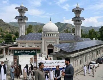 A mosque in Khyber Pakhtunkhwa with solar panels installed on its roof and minarets amidst news coverage and local observers.