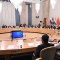A wide-angle photograph capturing a formal, high-level diplomatic conference in a grand, ornate room. In the immediate foreground, two officials in traditional Afghan attire (shalwar kameez and black turbans) are seated with their backs to the camera at a large, circular wooden conference table. Around the table, dozens of other delegates in suits are seated. In the background, mounted on the wall, are the flags of Russia, Afghanistan (current Taliban flag), India, China, Kyrgyzstan, Tajikistan, Turkmenistan, and Uzbekistan. A central audio-visual unit sits in the middle of the table. The room has large chandeliers and classical decor.