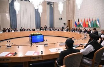 A wide-angle photograph capturing a formal, high-level diplomatic conference in a grand, ornate room. In the immediate foreground, two officials in traditional Afghan attire (shalwar kameez and black turbans) are seated with their backs to the camera at a large, circular wooden conference table. Around the table, dozens of other delegates in suits are seated. In the background, mounted on the wall, are the flags of Russia, Afghanistan (current Taliban flag), India, China, Kyrgyzstan, Tajikistan, Turkmenistan, and Uzbekistan. A central audio-visual unit sits in the middle of the table. The room has large chandeliers and classical decor.