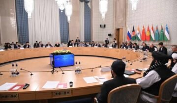 A wide-angle photograph capturing a formal, high-level diplomatic conference in a grand, ornate room. In the immediate foreground, two officials in traditional Afghan attire (shalwar kameez and black turbans) are seated with their backs to the camera at a large, circular wooden conference table. Around the table, dozens of other delegates in suits are seated. In the background, mounted on the wall, are the flags of Russia, Afghanistan (current Taliban flag), India, China, Kyrgyzstan, Tajikistan, Turkmenistan, and Uzbekistan. A central audio-visual unit sits in the middle of the table. The room has large chandeliers and classical decor.