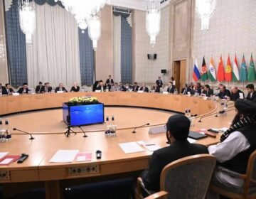 A wide-angle photograph capturing a formal, high-level diplomatic conference in a grand, ornate room. In the immediate foreground, two officials in traditional Afghan attire (shalwar kameez and black turbans) are seated with their backs to the camera at a large, circular wooden conference table. Around the table, dozens of other delegates in suits are seated. In the background, mounted on the wall, are the flags of Russia, Afghanistan (current Taliban flag), India, China, Kyrgyzstan, Tajikistan, Turkmenistan, and Uzbekistan. A central audio-visual unit sits in the middle of the table. The room has large chandeliers and classical decor.