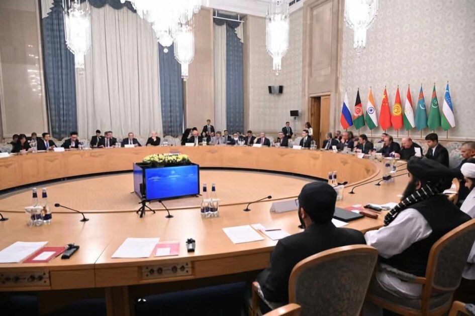 A wide-angle photograph capturing a formal, high-level diplomatic conference in a grand, ornate room. In the immediate foreground, two officials in traditional Afghan attire (shalwar kameez and black turbans) are seated with their backs to the camera at a large, circular wooden conference table. Around the table, dozens of other delegates in suits are seated. In the background, mounted on the wall, are the flags of Russia, Afghanistan (current Taliban flag), India, China, Kyrgyzstan, Tajikistan, Turkmenistan, and Uzbekistan. A central audio-visual unit sits in the middle of the table. The room has large chandeliers and classical decor.