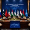 Empty diplomatic conference hall prepared for the 8th Moscow Format Consultation on Afghanistan, with Pakistan, Russia, China, Iran, Tajikistan, Uzbekistan, the Islamic Emirate of Afghanistan and other participating flags displayed behind vacant chairs.